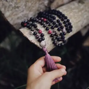 Home A close-up image of a hand holding a beaded necklace with a tassel, resting on a log outdoors.