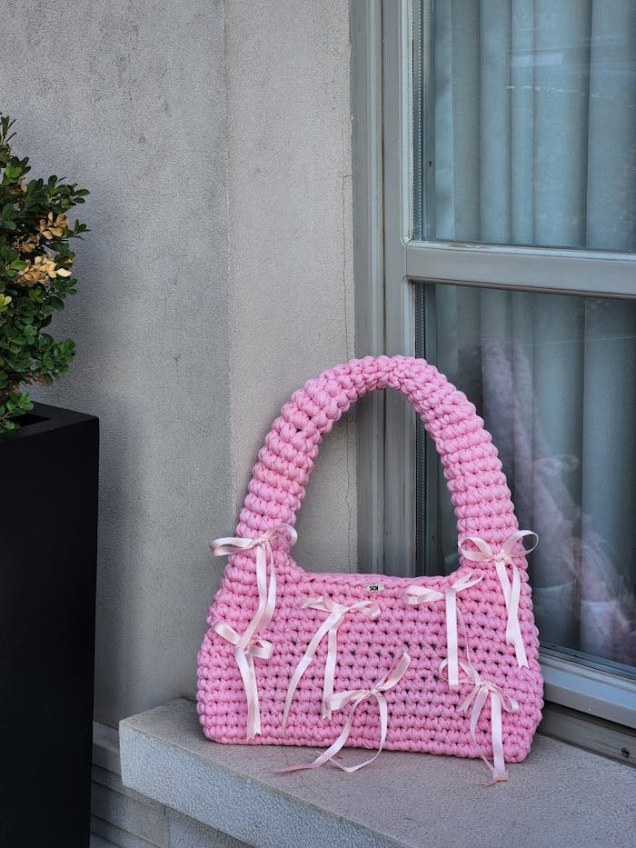 Fashionable pink knitted bag with ribbons displayed on a windowsill, showcasing chic design.